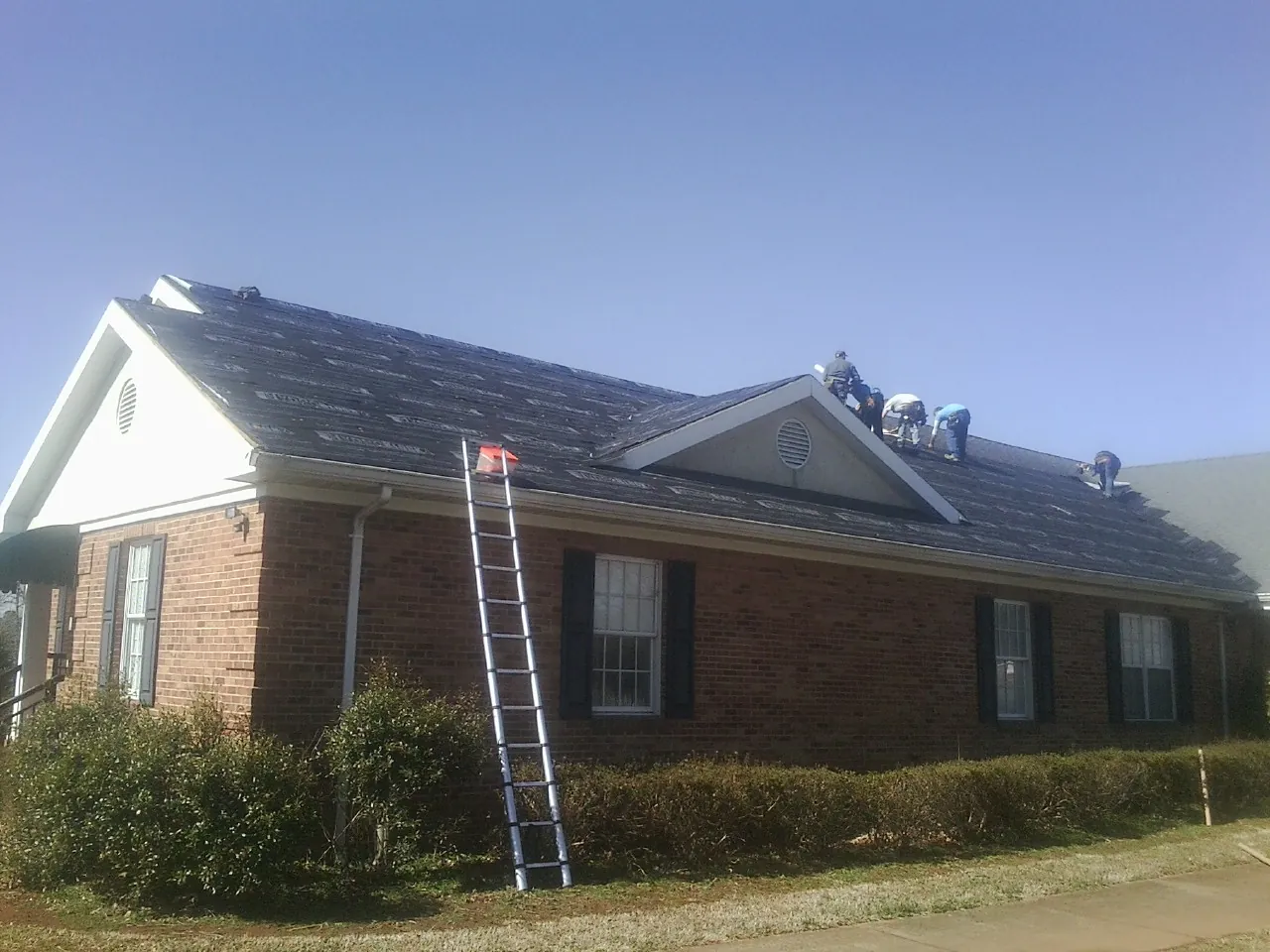 Crew tearing shingles off a brick ranch roof with a ladder set — Emergency Roof Repair work by our crew in Arverne