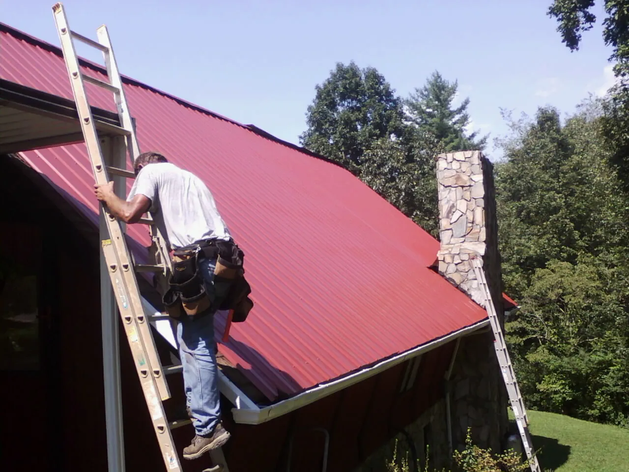 Roofer on a ladder beside a red metal roof and stone chimney — Soffit & Fascia Repair by Roofing Brothers in Arverne